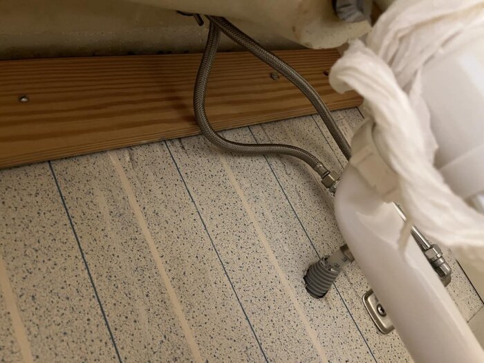 Rör och slangar under diskbänk med träregel och kaklat vägg. Vattenavstängning verkar saknas i detta utrymme.
