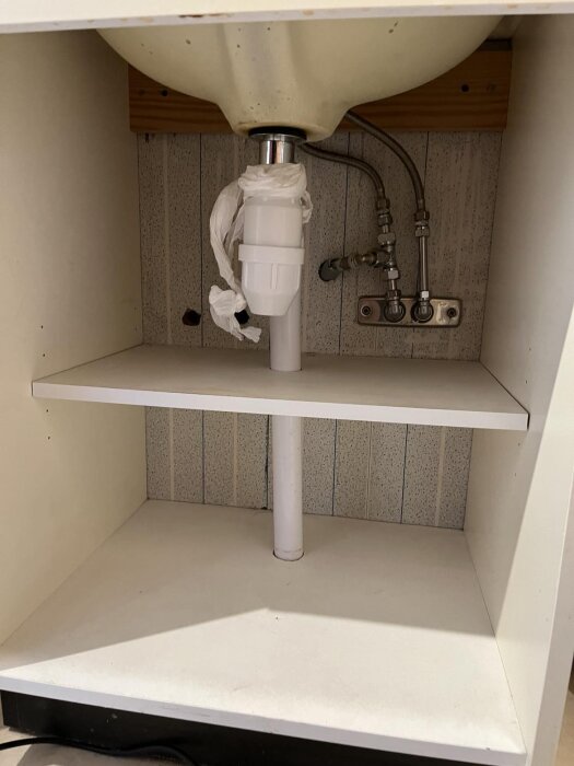 Under ett diskhon med synliga rör och en hylla, med plastpåse kring vattenlås.