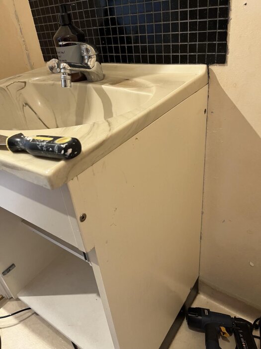 Tvättställ i badrum med vitt underskåp, skruvmejsel och flaska vid kranen; vattenavstängning saknas i rummet.
