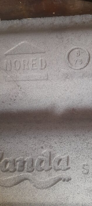 Betongplatta med inskriptionerna "NORED" inom en ram och "Kamela" i kursiv stil, samt siffrorna "8" och "75" inuti en cirkel.