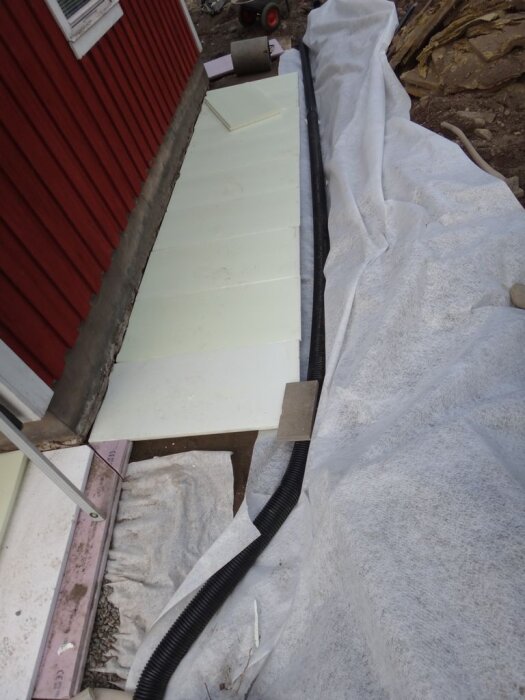 Dräneringsarbete vid husgrund med dräneringsrör, markisolering och fiberduk synliga vid en röd husvägg.