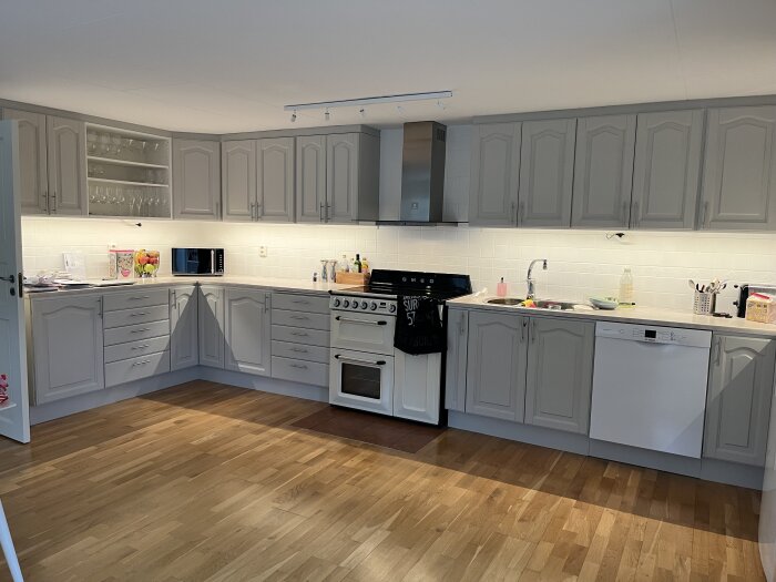 Kök med grå överskåp och diskbänk, LED-belysning under skåpen, spis och diskho synliga, samt träparkettgolv i ett modernt utförande.