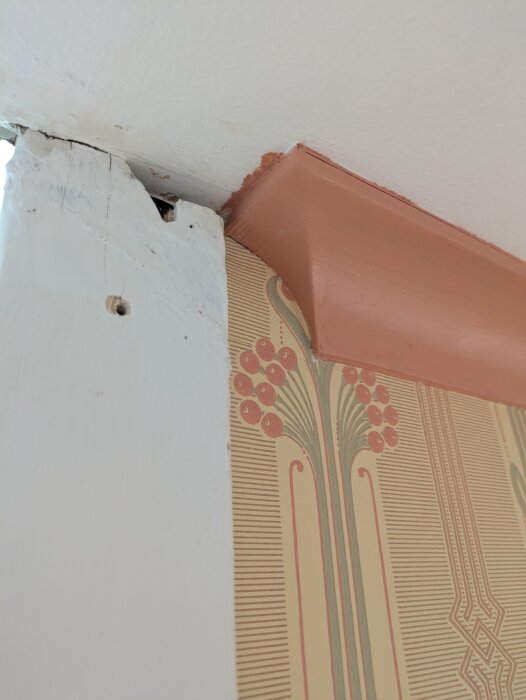 Nära bild på ett möte mellan vägglister med en orange målning och tapet med art déco-mönster, visar maskering och en orenoverad del av väggen.