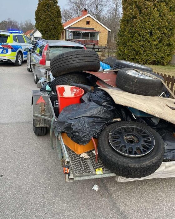 Släpvagn full med däck och byggmaterial, parkerad bakom en bil, med en polisbil i närheten. Hus och träd syns i bakgrunden.