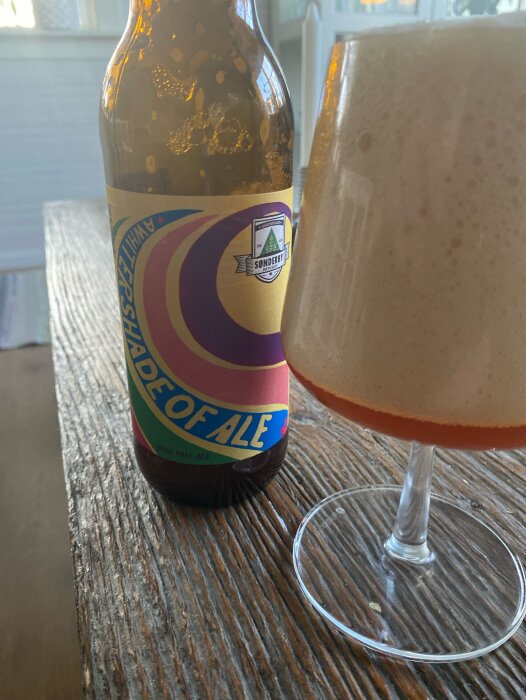 Flaska med etiketten "Amity Ale" och ölglas med rikligt skum på ett träbord.