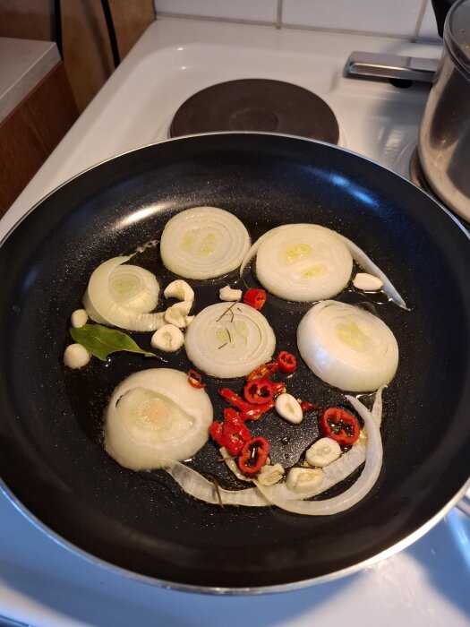 Ringskivor av lök, skivad chili, vitlök och ett lagerblad steks i olja i en stekpanna på en spis.