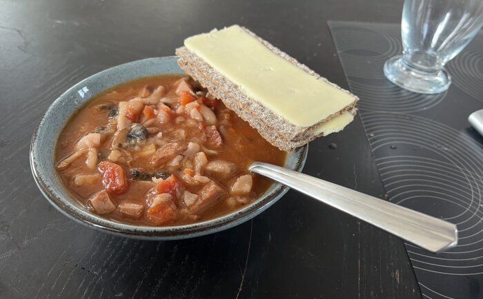 Skål med korvsoppa och knäckebröd med ost på ett bord, bredvid ett glas och en sked.