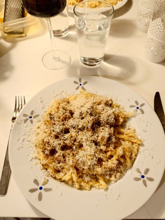 Kycklingpasta på en tallrik, toppad med basilika och mycket riven parmesan, serverad med ett glas rött vin och vatten på ett dukat bord.