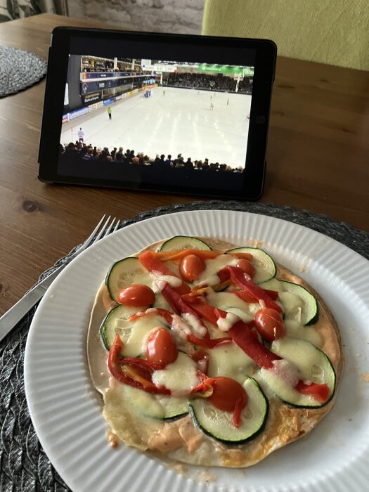 Tortillapizza med zucchini, tomater, grillad paprika och mozzarella framför surfplatta som visar en bandymatch.