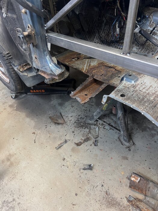 Bilrenovering pågår med rostiga delar på golvet, en domkraft och synliga exponerade metalldelar i karossen.