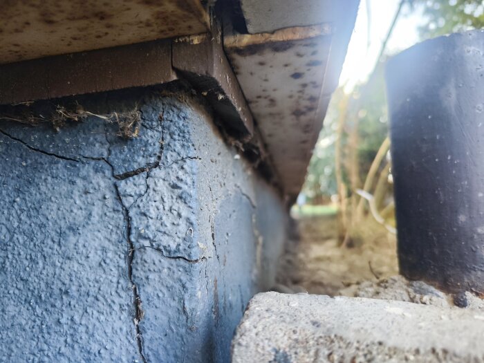Sprucken husgrund med rostig balk ovanför, omgiven av jord och ett svart rör.