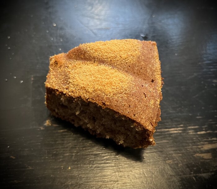 En bit pepparkakskaka bakad med mandelmjöl och vetemjöl, med ojämn ströbrödstoppning, ligger på ett mörkt underlag.