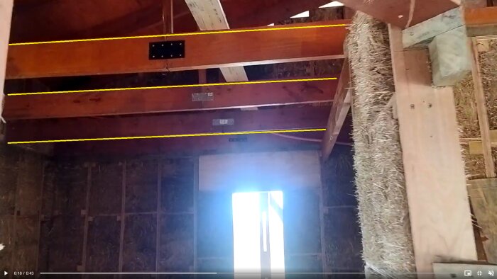 Interior view of a building under construction with visible wooden ceiling beams and a metal connector plate, alongside a window emitting bright light.