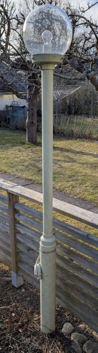 Gammal lyktstolpe från 1950-talet med glaskupol, stående i trädgård intill trästaket.