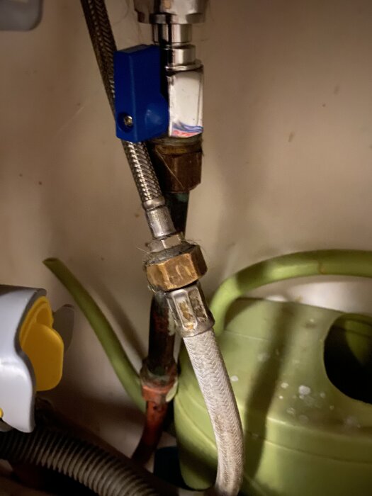 Diskmaskinens vattenslang ansluten till en ventil med Aquastop-funktion, tillsammans med andra rör och en grön vattenkanna i bakgrunden.