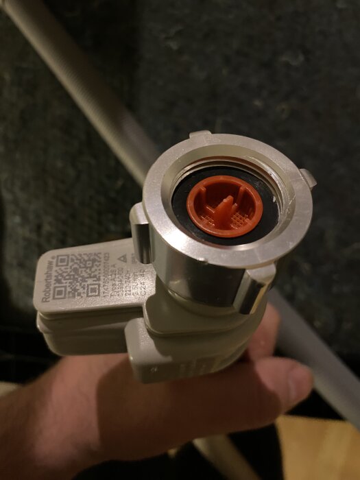 Grå och orange Aquastop-anslutning med QR-kod hålls i hand, slangen i bakgrunden. Läggs upp med fråga om diskmaskinsanslutning och adapter.