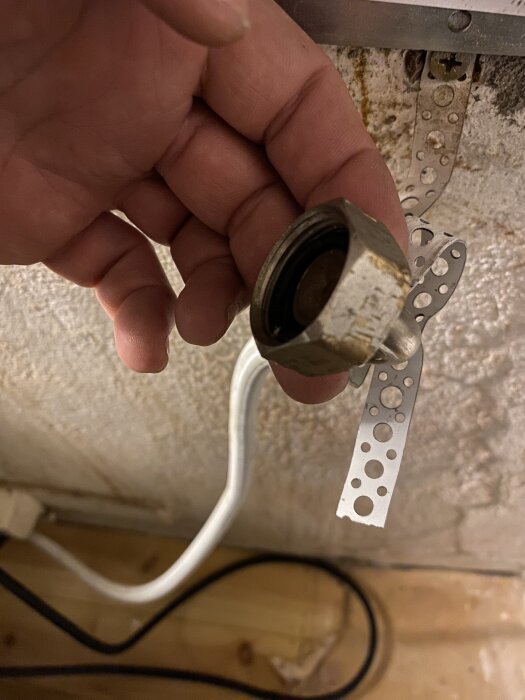 Hand håller en metallanslutning för diskmaskinens vattenledning, med en vit slang och perforerad metallremsa i bakgrunden.
