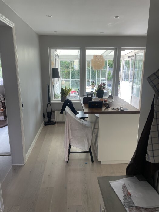 Ljust kök med utsikt mot ett uterum, visande en grå stol med tyg över ryggen vid diskbänken och stora fönster med mycket ljusinsläpp.