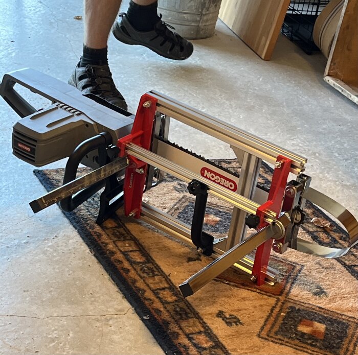 En batteridriven motorsåg från Jula/Meec Tools med ett Oregon sågverk, placerad på ett mönstrat golv tillsammans med en fot i sandaler.