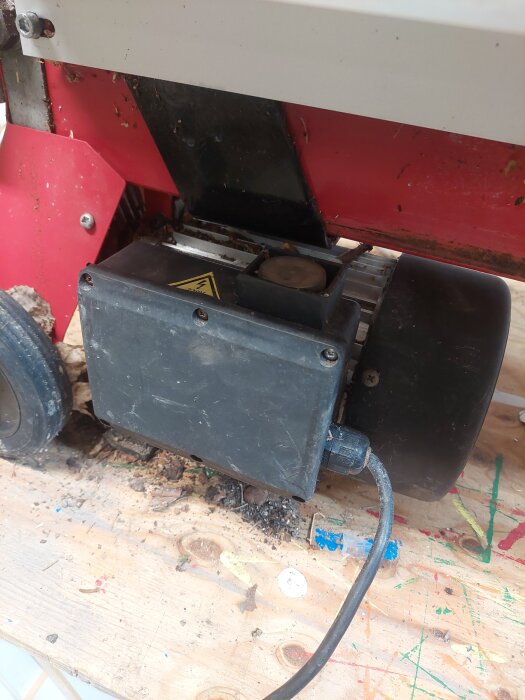 En vedklyvs motor och elektrisk kopplingsbox på en arbetsbänk. Sladdar och ett varningsmärke är synliga på motorhöljet.