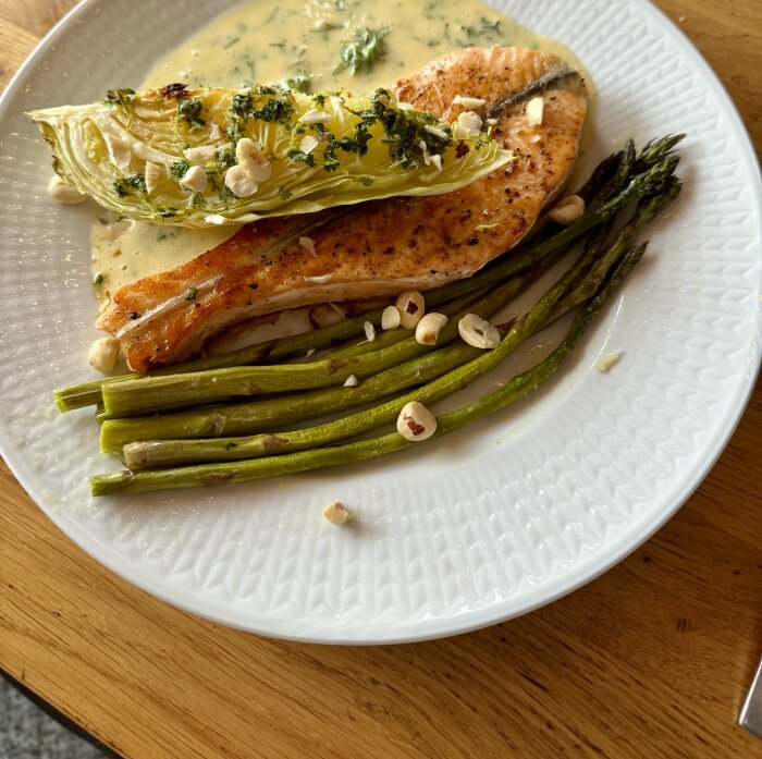 Tallrik med laxfilé, spetskål, sparris och örtsås toppad med nötter på ett bord.