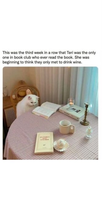Vit katt vid bord med uppslagen bok, kaffekopp och ljusstake, ser ut att delta i bokklubbsmöte.