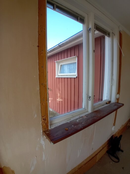Gammal fönsterbräda i trä monterad under ett fönster med utblick mot en röd byggnad. Brädan verkar ha släppt något från väggen.
