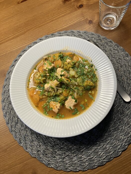 Laxsoppa i en vit tallrik på ett bord, garnerad med örter, placerad på en grå bordstablett.