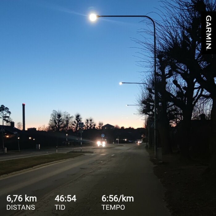 Kvällsvy över en upplyst vägkant med gatubelysning och bilar i bakgrunden under en blå skymningshimmel, text med distans och tempo från Garmin.