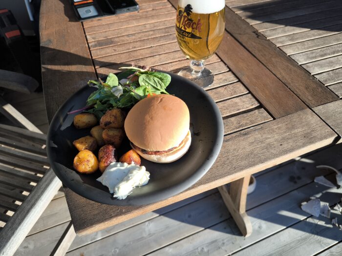 Grillad hamburgare med helstekt potatis, sallad och bearnaisesås på en tallrik, samt en ölglas på ett träbord i solljus.