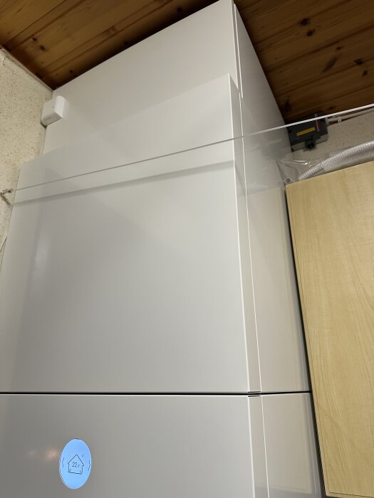 Vitt överskåp i tvättstuga, stående nära taket, med digital display som visar 22°C. Skåp monterat på vita fötter, lämnar 3 cm till taket.