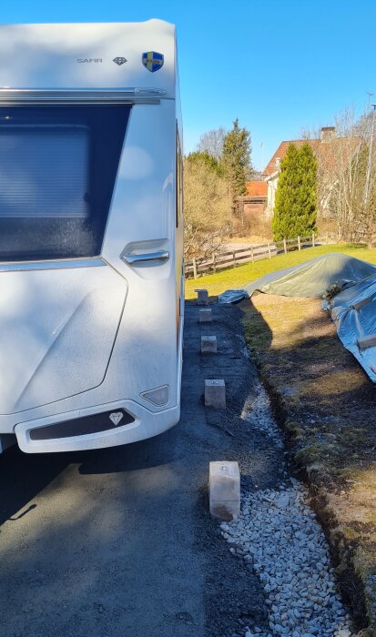 Husvagn backad på plats nära kanten av en smal uppfart, omgiven av stenblock, med grönt och ett hus i bakgrunden.