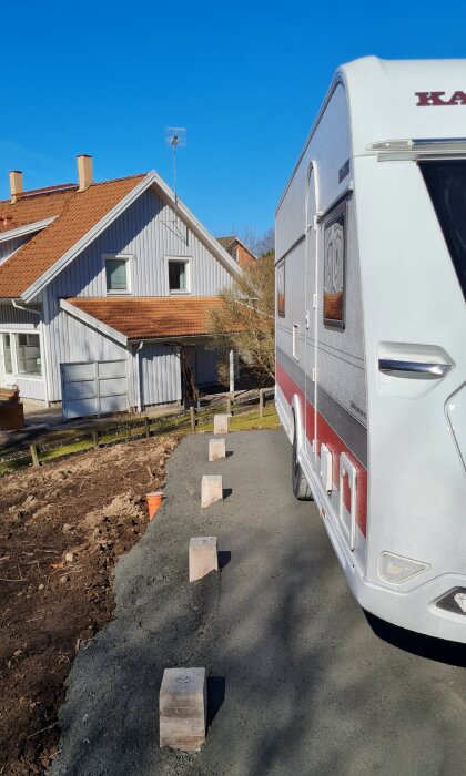 Husvagn parkerad på en asfalterad uppfart bredvid ett hus, med träklossar längs kanten för att markera garageplatsen.
