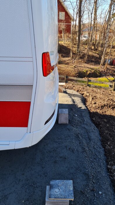 Tail light of a white caravan parked on gravel near a house, bordered by a wooden fence and trees in the background.
