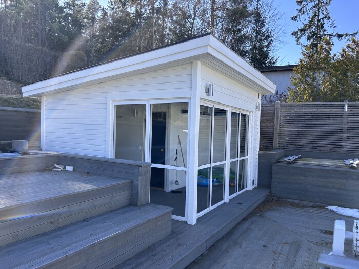 Vit byggnad med stora glasdörrar, omgiven av trädäck och träpanel, troligen en bastu.
