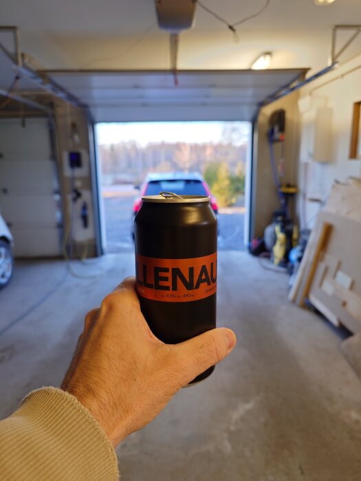 En hand håller i en ölburk i en garageuppfart med en central bil i bakgrunden.