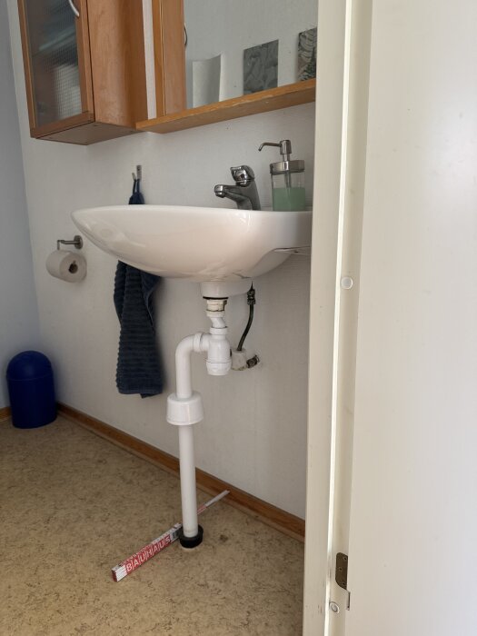 Handfat med synligt avloppsrör i en liten gästtoalett. Vänster om handfatet finns en handduk och en toalettpappershållare.