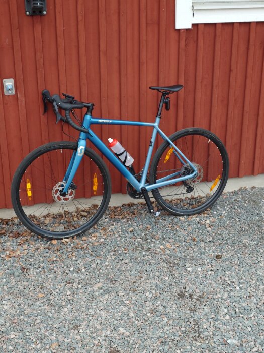 Blå cykel lutad mot en röd trävägg, stående på en grusad yta, med en vattenflaska monterad på ramen.