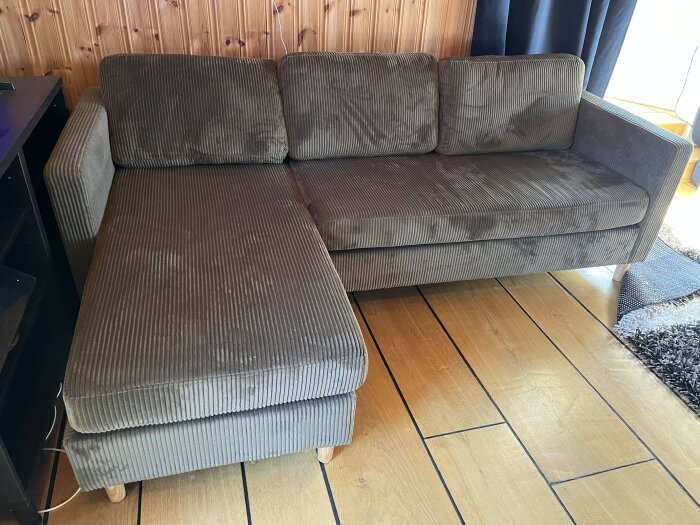 Olivgrön soffa med schäslong, placerad på ett trägolv bredvid en svartmöbel, framför en träpanelvägg med gardiner till höger.