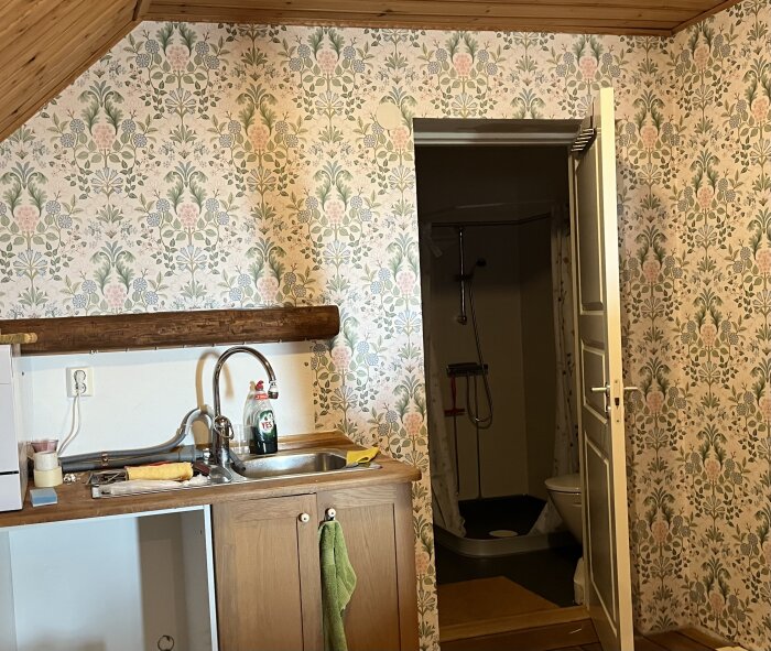 Blommig tapet i ett rum med snedtak, diskho, och dörr som leder till ett badrum. Verktyg och rengöringsmedel syns på bänken.