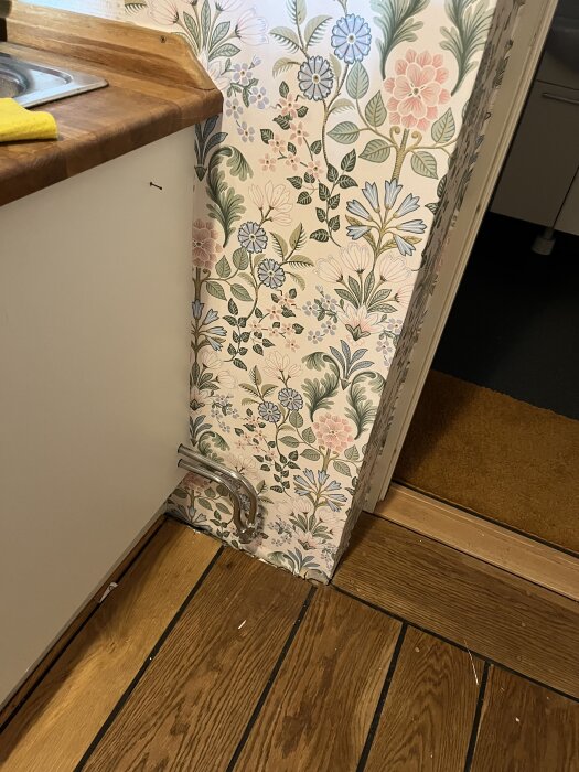 Blommig tapet vid rör i kök, med synlig trälaminat på golvet.