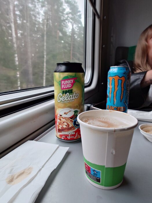 Alkoholfria drycker och en kaffekopp på ett tågbord med utsikt över skog genom fönstret.
