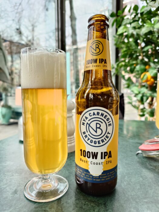 En glasflaska med etiketten "100W IPA West Coast IPA" bredvid ett glas fyllt med gyllene öl på ett bord.
