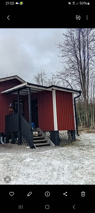 Röd stuga med takutbyggnad och stupande takränna, omgiven av träd i vinterlandskap. Snö täcker marken lätt.