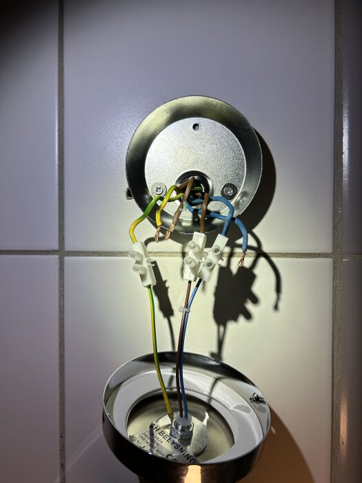 Elinstallation av lampa med synliga kablar: en gul/grön jordad, en brun fas och en blå neutral, monterade på kakel i ett badrum.