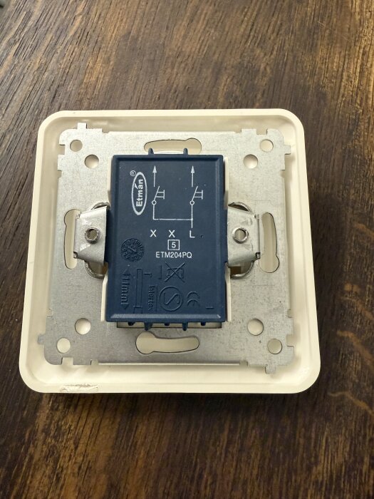 Elektrisk brytare med etiketten "Eltman ETM204PQ," monterad i en metallram på en träram. Brytaren har symboler och terminaler markerade.