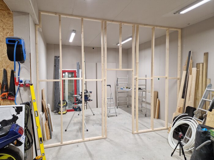 Skiljevägg under uppbyggnad i ett garage med träreglar, verktyg, stegar och byggmaterial runtomkring i rummet.