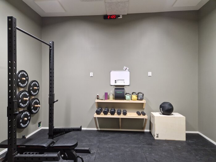 Hemmagym med skivstångsställning, viktskivor på väggen och en hylla med kettlebells, hantlar och ett jumping box. Vit skrivtavla i bakgrunden.