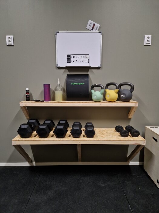 Hylla med vikter och kettlebells av olika storlekar, en whiteboard ovanför, del av ett hemmagym.