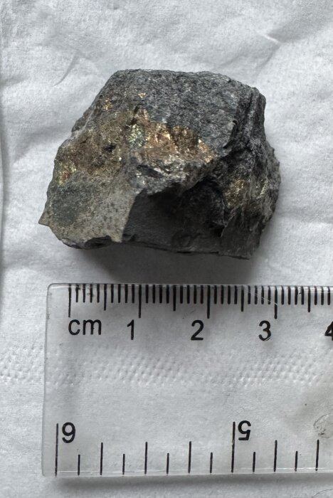 En glittrande sten med skimrande lager, eventuellt kvarts eller glimmerskiffer, ligger bredvid en linjal som visar dess storlek i centimeter.